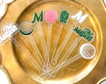 May include: A set of seven clear acrylic drink stirrers with various designs, including golf balls, the words "The Masters", and a green "M".