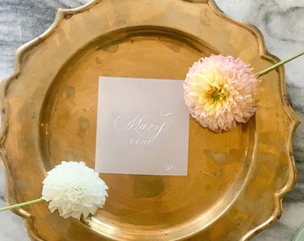 May include: A gold charger plate with a white card that says "Mary Cook" on it. There are two pink and white flowers on the plate.