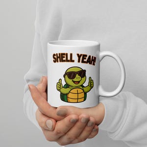 Könnte beinhalten: Eine weiße Keramiktasse mit einer grünen Schildkröte, die eine Sonnenbrille trägt und den Daumen nach oben zeigt. Auf der Tasse steht der Text "Shell Yeah!" gedruckt.