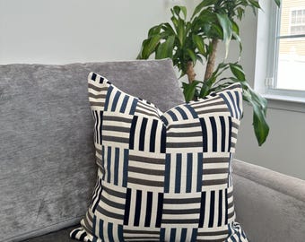 Puede incluir: Cojín decorativo cuadrado con un patrón geométrico en azul marino, gris y blanco. El cojín está colocado sobre un sofá gris. Una planta verde es visible en el fondo.
