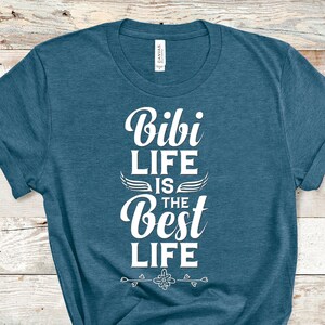 May include: A teal blue t-shirt with the text "Bibi Life is the Best Life" in white. The text is surrounded by a decorative design with wings and hearts.