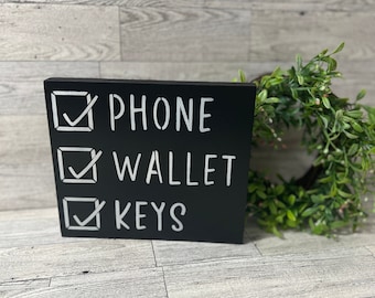 May include: Black wooden sign with white checkboxes and text. The checkboxes are checked and the text reads "PHONE", "WALLET", and "KEYS".