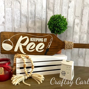 May include: Wooden paddle sign with the text "Keeping it Ree" in white paint. The sign has a fishing bobber and fishing rod painted on it. The sign is sitting on a white crate with a small white house-shaped candle holder that says "Home Sweet Home".