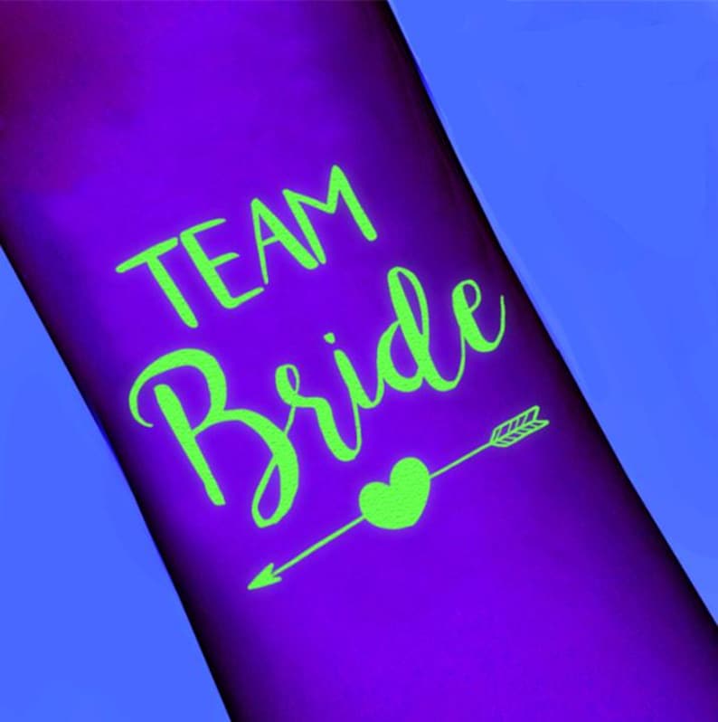 May include: A purple surface with the words "TEAM Bride" in bright green script. A green heart is pierced by an arrow, also in green. The background is a gradient of purple and blue.
