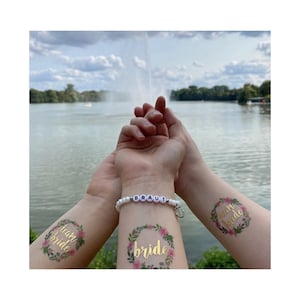Könnte beinhalten: Drei Arme mit temporären Tattoos und einem Armband. Die Tattoos zeigen Blumenkränze mit den Worten "bride" und "team bride" in goldfarbenen Buchstaben. Ein Perlenarmband schreibt "Braut". Der Hintergrund zeigt einen See und Bäume.
