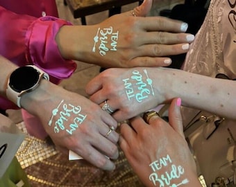 Op de afbeelding: Close-up van handen met tijdelijke tatoeages met de tekst "Team Bride" in een lichtblauw scriptlettertype, met een pijl- en hartontwerp. Sommige handen hebben ringen en een horloge is zichtbaar. Het beeld suggereert een bruidsfeest.