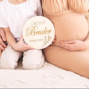 May include: A round wooden sign with the words "GROSSER Bruder FEBRUAR 2023" in gold lettering, held by a child. The sign has a floral design and is held in front of a pregnant person.
