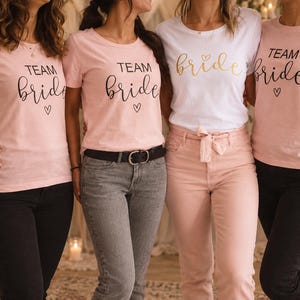 Könnte beinhalten: Drei hellrosa T-Shirts und ein weißes T-Shirt mit dem schwarzen Text "TEAM bride" und einem kleinen Herzsymbol darunter. Die T-Shirts haben kurze Ärmel und einen Rundhalsausschnitt. Der Text ist in einer Kombination aus Block- und Schreibschrift gedruckt.