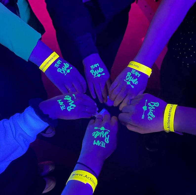 May include: A group of hands with the words "Team Bride" and a heart-shaped arrow design glowing in neon green. Yellow wristbands with the text "Actionarena.info" and "www.Action" are also visible. The scene is illuminated with a purple glow.