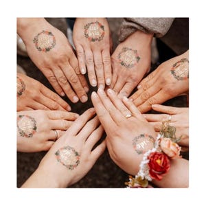 Könnte beinhalten: Mehrere Hände mit temporären Tattoos mit den Worten "Team Bride" und "Bride" in floralen Kränzen. Eine Hand zeigt ein florales Handgelenk-Korsett mit roten und rosa Rosen. Die Hände sind kreisförmig angeordnet.
