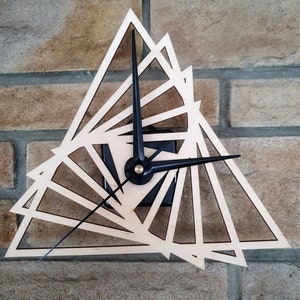 Può includere: Un orologio in legno con lancette nere e un design geometrico di triangoli sovrapposti. L'orologio è montato su un muro di mattoni.