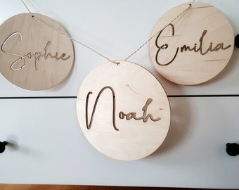 Peut inclure: Une guirlande en bois avec trois ornements circulaires en bois. Chaque ornement a un nom découpé dans une police cursive. Les noms sont "Sophie", "Émilia" et "Noé".