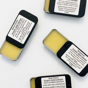 May include: Four rectangular metal tins containing yellow solid cologne, each with a black sliding lid featuring a white label. The labels list the scents Majestic Oud, Spiced Vanilla, Bergamot, and Sweet Tobacco, along with ingredients and 0.05oz weight.