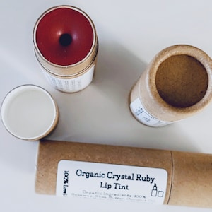 May include: An overhead shot of organic lip tint products. The image features a red lip tint in a cardboard tube, a separate empty tube, and a tube with a label that reads "Organic Crystal Ruby Lip Tint". The products are on a white surface.