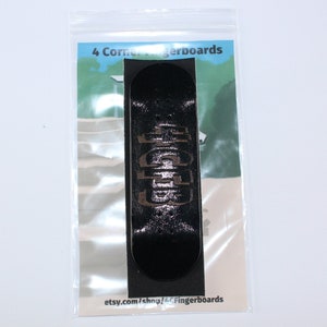 May include: Black fingerboard with gold lettering that reads "4 Corner Fingerboards" on a white background. The fingerboard is in a clear plastic bag.