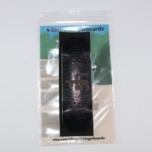 May include: A black fingerboard with a brown logo on it. The fingerboard is in a clear plastic bag with a white background that says "4 Corner Fingerboards" and "etsy.com/shop/4CornerFingerboards".
