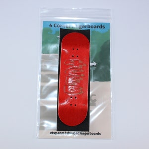 May include: A red fingerboard with a black and white graphic design. The fingerboard is in a clear plastic bag with a white background. The text "4 Corner Fingerboards" is printed on the bag. The text "etsy.com/shop/4CFingerboards" is printed on the bottom of the bag.