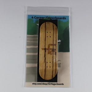 May include: A wooden fingerboard with a brown and black wood grain pattern. The fingerboard has a black grip tape and a logo that says "4 Corner Fingerboards".