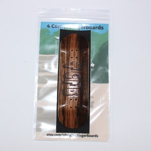 May include: A fingerboard made of wood with a dark brown and tan striped pattern. The fingerboard has a black graphic and is packaged in a clear plastic bag. The text "4 Corner Fingerboards" is visible.