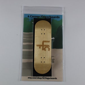 May include: A wooden fingerboard with the text "4 Corner Fingerboards" and "etsy.com/shop/4CFingerboards" printed on the packaging. The fingerboard is a light brown color and has a black graphic on it.