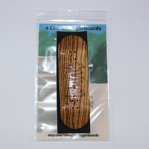 May include: A wooden fingerboard with a natural wood grain finish. The fingerboard is packaged in a clear plastic bag with a white and green background. The text "4 Corner Fingerboards" is printed on the bag. The text "etsy.com/shop/4cornerfingerboards" is printed on the bottom of the bag.