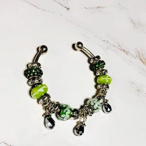 Könnte beinhalten: Ein silberfarbener Bauchnabelpiercing mit grünen und weißen Perlen und baumelnden schwarzen Tropfenanhängern. Die Perlen haben verschiedene Designs, darunter florale Muster und funkelnde grüne Akzente. Der Schmuck wird auf einer weißen Marmoroberfläche präsentiert.