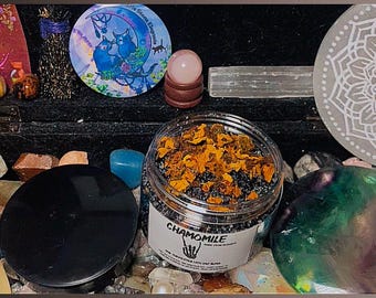 May include: A clear plastic jar filled with a dark gray granular substance topped with dried orange flower petals. The label on the jar features the text CHAMOMILE and a graphic of a skeletal hand. A black lid sits beside the open jar.