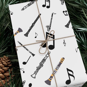 May include: A wrapped gift featuring a white background with a pattern of musical instruments and notes. The instruments include clarinets and saxophones in black and gray. The gift is tied with twine and rests on a bed of green pine needles and pine cones.