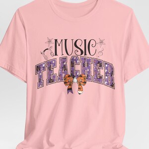May include: Pink t-shirt with the text "MUSIC TEACHER" in purple and black letters with a purple and orange bow. The text is surrounded by stars and musical notes.