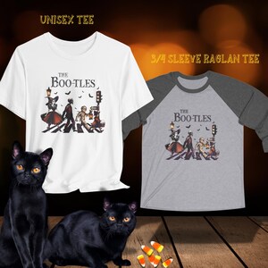 Op de afbeelding: Twee zwarte katten zitten op een houten oppervlak voor twee T-shirts. De T-shirts hebben een afbeelding van een Halloween-band die over een zebrapad loopt. De tekst op de T-shirts is "The Boo-tles".