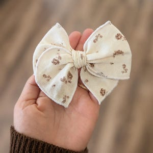 May include: A cream-colored fabric hair bow featuring a small, repeating brown floral pattern. The bow has a layered, multi-loop design with a central knotted fabric band and visible stitched edges, creating a soft, textured appearance.