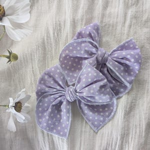 Op de afbeelding: Twee lavendelkleurige stoffen haarstrikken met witte stippen. De strikken zijn in het midden geknoopt en hebben een gestikte rand. De strikken liggen op een witte, getextureerde achtergrond met witte bloemen.