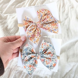 Puede incluir: Dos lazos para el pelo de tela floral sobre tarjetas. Los lazos presentan un colorido estampado de pequeñas flores en tonos naranja, rosa, azul y amarillo. Las tarjetas tienen el texto "PETAL + PINE BOWS". Los lazos están sujetos a una diadema elástica beige.