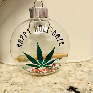 Könnte beinhalten: Transparente Glaskugel mit dem schwarzen Schriftzug "HAPPY HOLI-DAZE". Im Inneren befinden sich ein Marihuanablatt, eine gerollte Zigarette und bunte Konfetti. Die Kugel hat eine silberne Metallkappe.