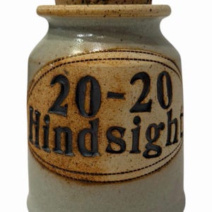 May include: A light grey ceramic jar with a cork stopper. The jar features a brown oval label with the words "20-20 hindsight" in black. The jar has a handmade, rustic aesthetic.