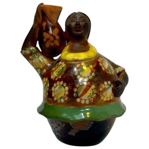 Puede incluir: Figura de cerámica de una mujer que lleva una jarra. La figura tiene una tez marrón oscura, un cuello amarillo y un vestido con estampados florales en amarillo, verde y rojo. La base es negra con ribete verde.