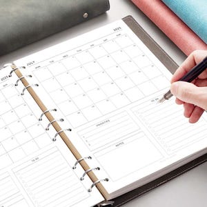 May include: A white planner with a black cover and silver rings. The planner has a monthly calendar for July 2025, a to-do list, a priorities section, and a notes section.