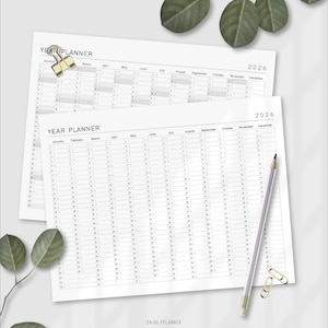 May include: Two white 2026 year planner sheets with the text "YEAR PLANNER" and monthly calendars. A pencil, paper clips, and decorative leaves are on the surface. The bottom right corner has the text "ENJULYPLANNER".