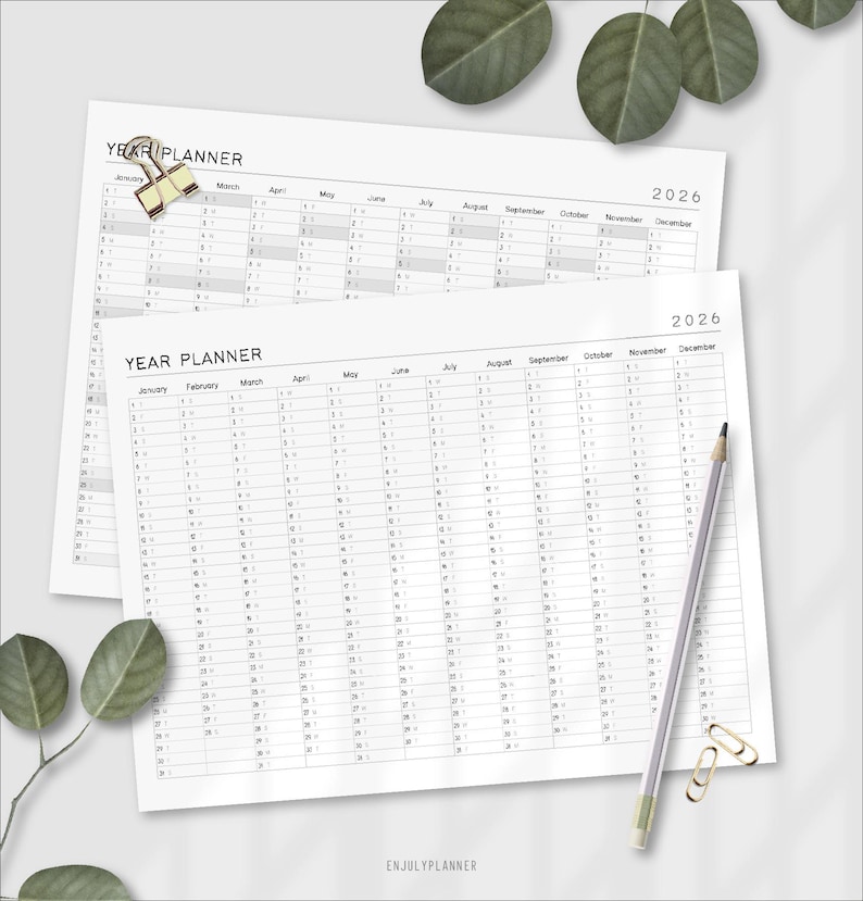 May include: Two white 2026 year planner sheets with the text "YEAR PLANNER" and monthly calendars. A pencil, paper clips, and decorative leaves are on the surface. The bottom right corner has the text "ENJULYPLANNER".