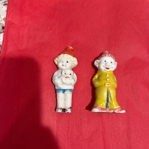 May include: Two small ceramic figurines. The left figurine is a child in a red hat holding a white dog. The right figurine is a dwarf wearing a yellow coat and a red hat with the word Dopey printed on the front in white capital letters.