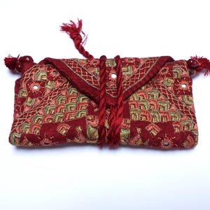 May include: An embroidered clutch bag in red, pink, and gold. The fabric has a geometric pattern with small mirrors. The bag has a flap closure secured with a red cord and tassels. A decorative, ethnic-inspired accessory, ideal for carrying essentials.
