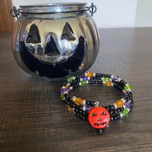 May include: A black beaded bracelet with orange, green, and purple accents. A small orange pumpkin charm with a black face is attached to the bracelet.