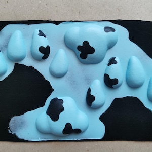 May include: A blue fabric patch with a cow print design. The patch has a raised, textured surface with black spots on a light blue background.