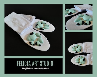 May include: A pair of white socks with a light blue paw print design. The paw prints have black spots. The socks are folded and displayed on a black background. The text "FELICIA ART STUDIO" and "Etsy/Felicia art studio shop" is visible at the bottom of the image.
