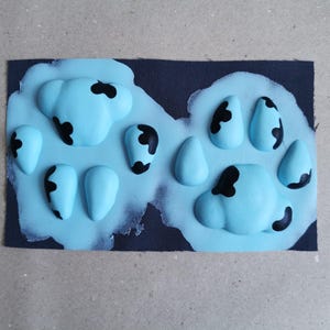May include: A close-up of a blue paw print with black markings on a dark fabric background. The paw print is made of a soft, pliable material.