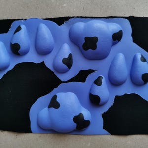 May include: A close-up of a blue fabric patch with black spots, resembling a cow print. The patch is made of a soft, textured material and has a raised, 3D design.