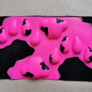 May include: A pink fabric patch with black painted spots in a cow print pattern. The fabric is textured and has a raised design.