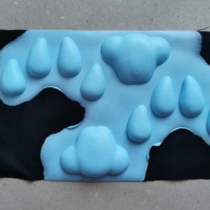 May include: A blue fabric patch with raised paw prints and cloud shapes. The patch is sewn onto a black fabric background.