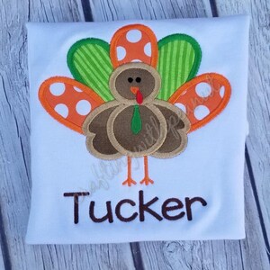 May include: White t-shirt featuring a colorful turkey design with orange, green, and brown accents. The turkey has a red wattle and a green tie. The name "Tucker" is printed in brown letters below the turkey. A festive apparel item.