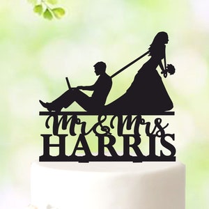 Könnte beinhalten: Schwarze Silhouette Tortenaufsatz, der eine Braut zeigt, die einen Bräutigam zieht, der sich auf einen Laptop konzentriert. Der Aufsatz trägt die Aufschrift "Mr & Mrs Harris", passend für eine Hochzeitstorte.